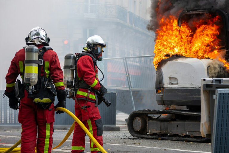 Zwei Feuerwehrleute mit Schläuchen neben einem brennenden Bagger auf einer abgesperrten Strasse