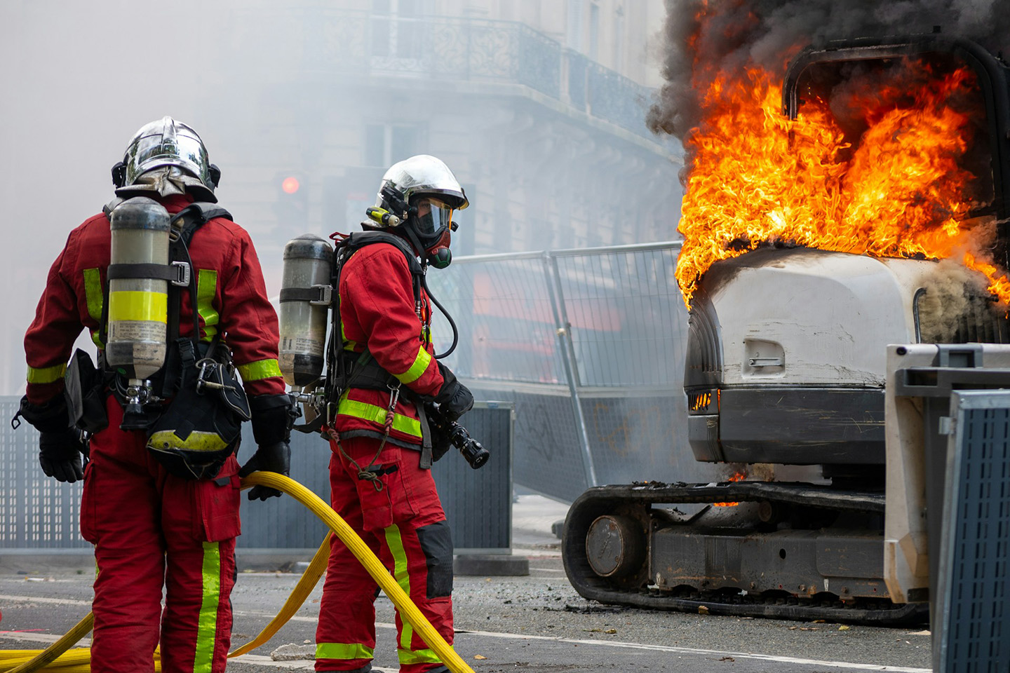 Zwei Feuerwehrleute mit Schläuchen neben einem brennenden Bagger auf einer abgesperrten Strasse