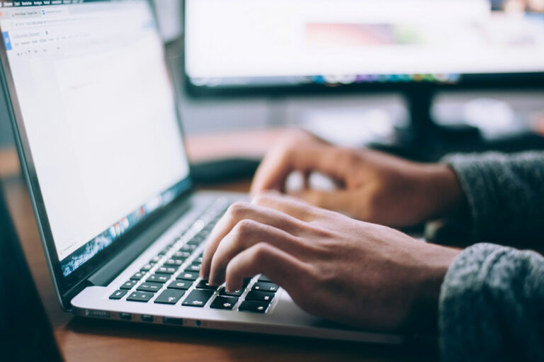 Arbeitssituation an einem geöffneten Laptop, zwei Hände tippen auf der Tastatur, im Hintergrund ist ein Teil eines PC-Monitors zu sehen